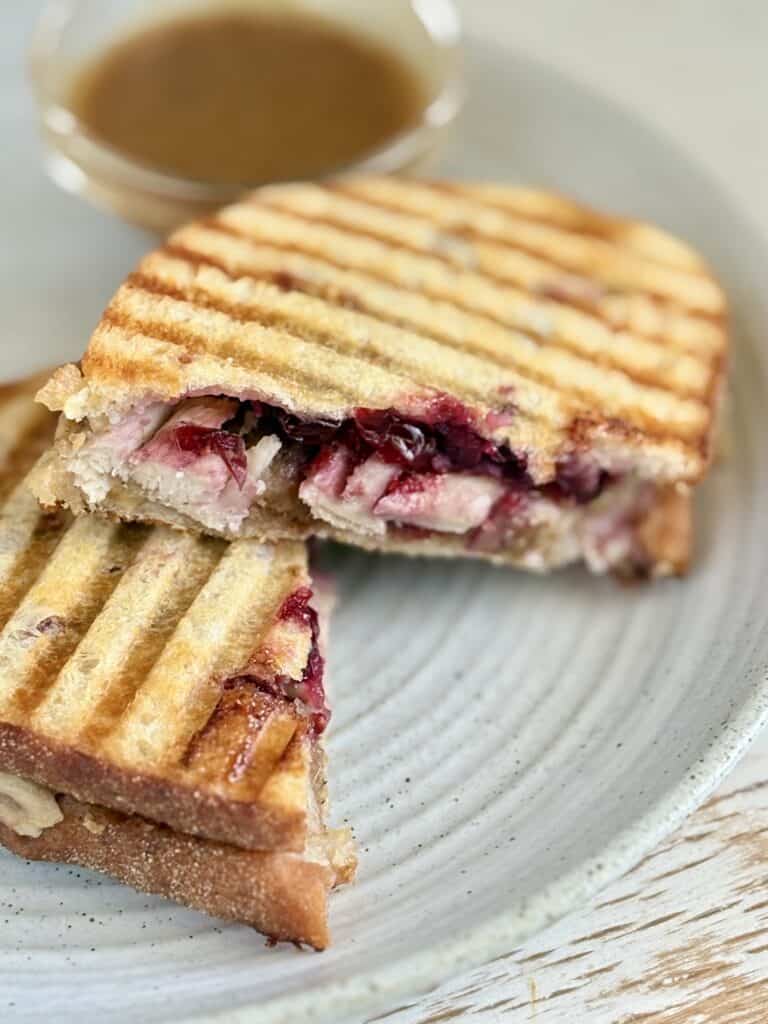 Close-up of a grilled turkey and cranberry panini with grill marks, served with a side of dipping sauce on a white plate.