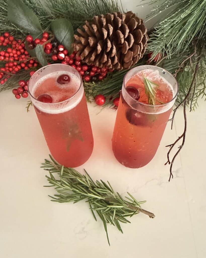Two glasses of cranberry pomegranate sparkling drink garnished with cranberries and mint, surrounded by holiday greenery and pinecones.