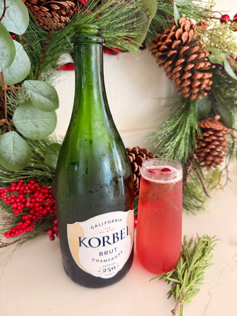 Bottle of Korbel Brut Champagne with a glass of cranberry pomegranate sparkling drink, surrounded by festive holiday greenery and pinecones.