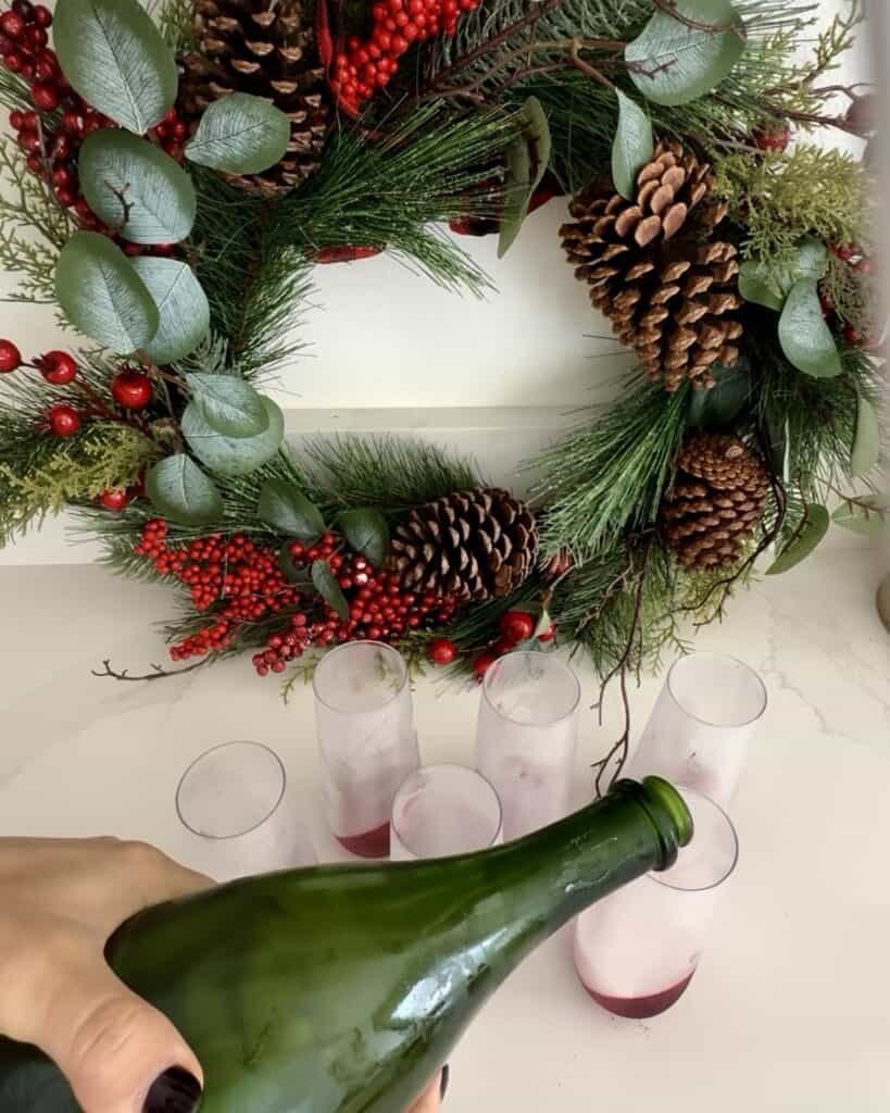 Festive holiday wreath with pinecones, red berries, and greenery, with glasses of cranberry pomegranate sparkling drink on a white table.