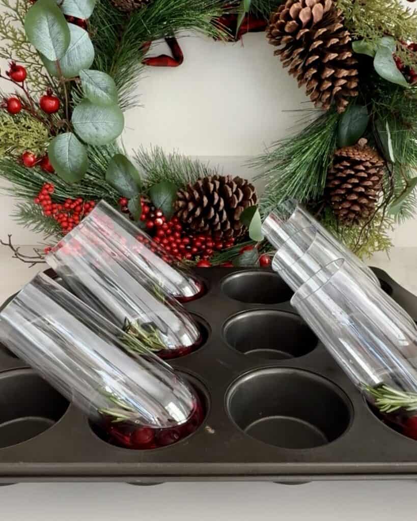 Festive holiday wreath with pinecones, red berries, and greenery, with glasses of cranberry pomegranate sparkling drink on a black tray.