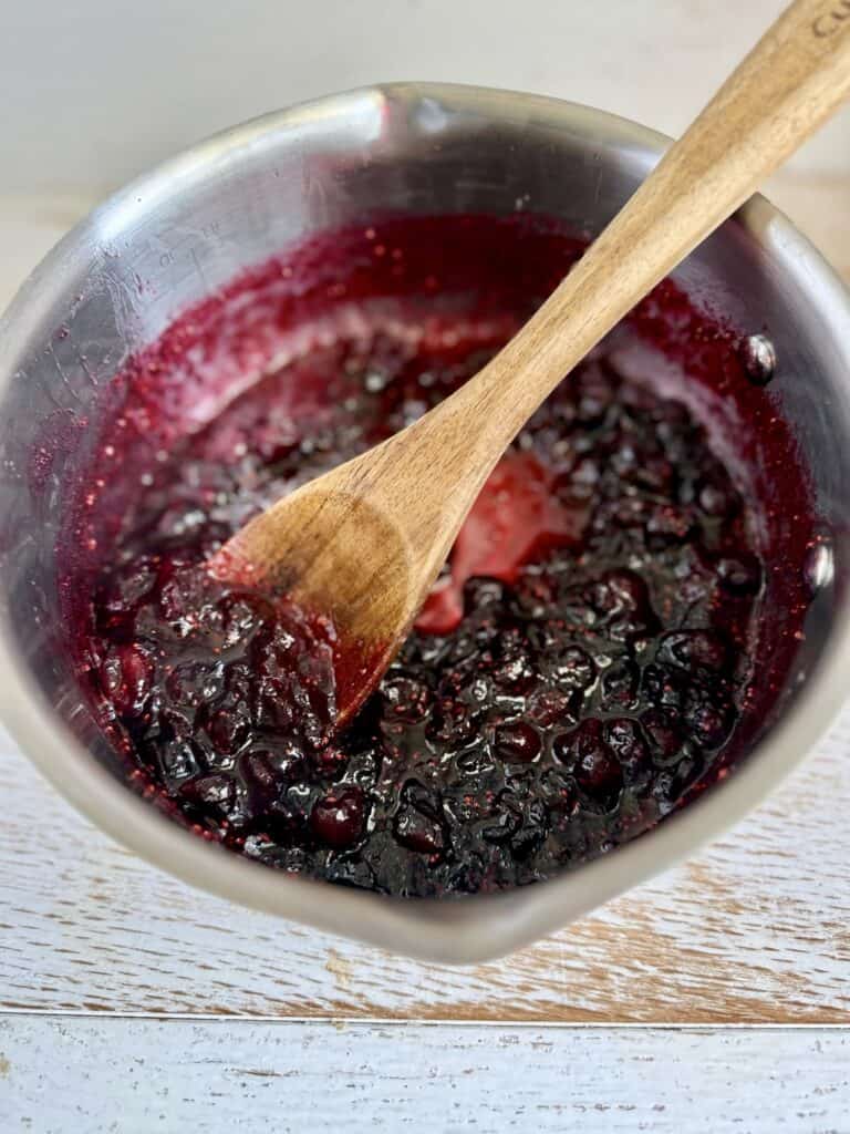 Close-up of homemade cranberry sauce in a silver pot with a wooden spoon, showcasing a rich, dark purple color and textured surface.