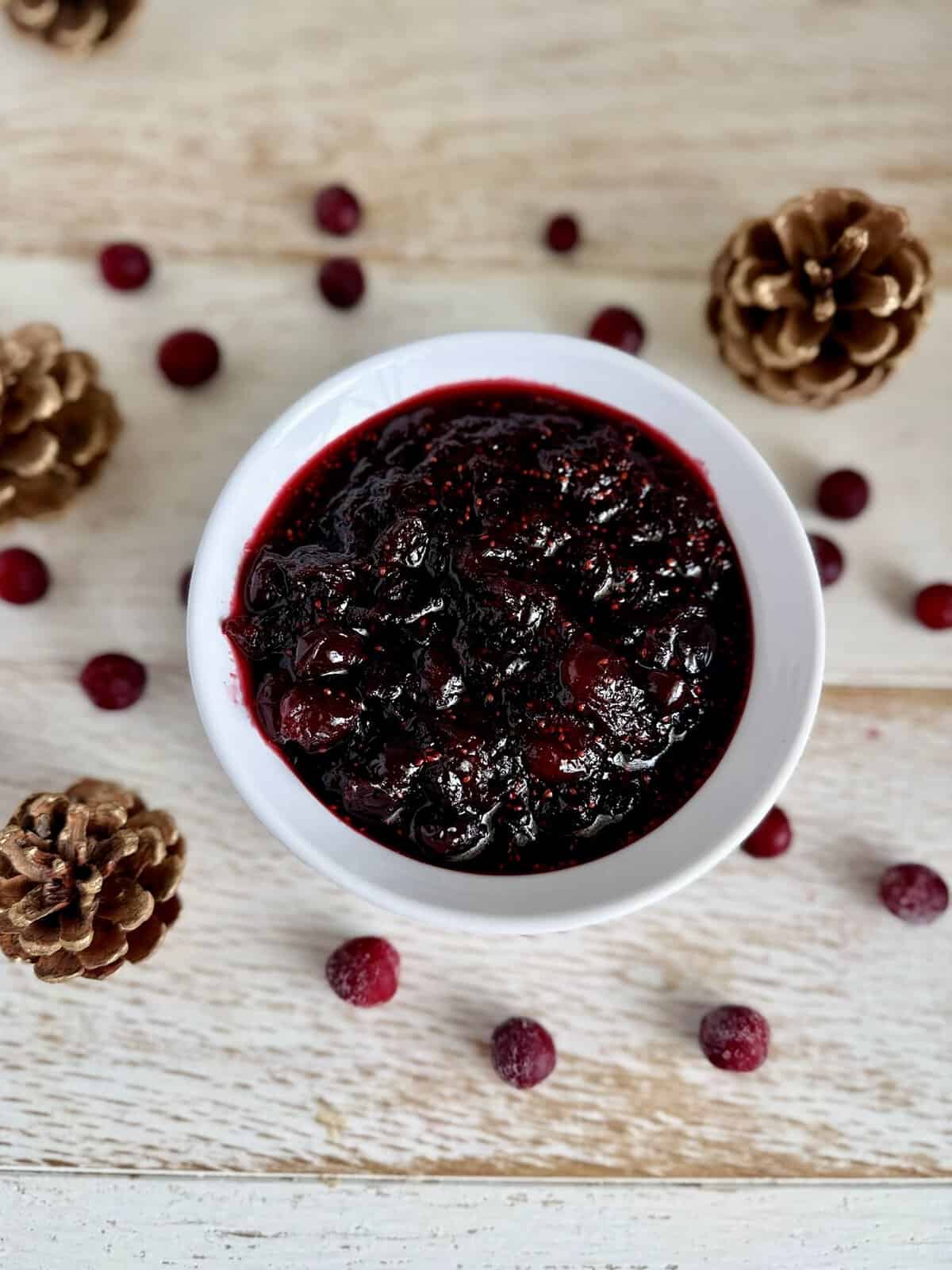 Close-up of a bowl of homemade cranberry sauce surrounded by pinecones and fresh cranberries on a wooden surface.